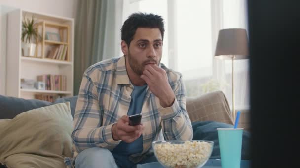 Średni Steadicam młodego człowieka rasy mieszanej siedzi na kanapie w salonie w domu, jedząc popcorn, oglądając telewizję, klikając kanały z pilotem, patrząc zainteresowany - Materiał filmowy, wideo