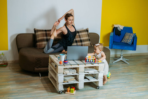 Młoda kobieta matka siedzieć na kanapie ze swoim malutkim córeczką w domu robi ćwiczenia jogi, oglądając online zajęcia jogi na laptopie. Sportowa kobieta z dzieckiem ćwiczą w salonie - Zdjęcie, obraz