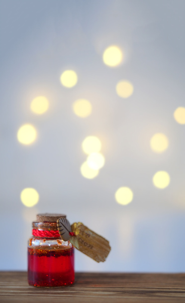 A small glass jar with red-gold liquid and a gold tag stands on a wooden table on a blur of a table with a side from a garland, a vertical image with a soft focus and a place for text - Photo, Image