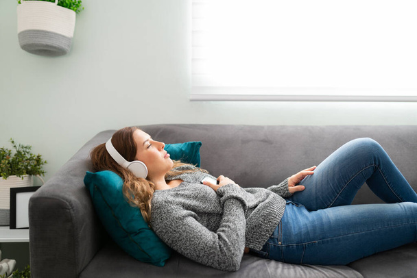 Vista lateral de la joven hermosa mujer tumbada en el sofá, relajándose en su sala de estar y escuchando música con auriculares - Foto, Imagen