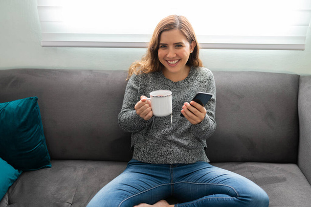 Портрет улыбающейся молодой женщины, сидящей дома на диване с чашкой горячего чая и смартфоном - Фото, изображение