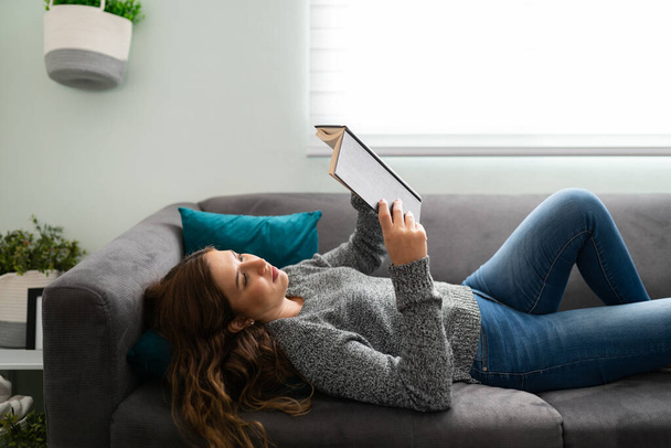 Vista lateral de una hermosa joven acostada en el sofá y leyendo un libro  - Foto, imagen