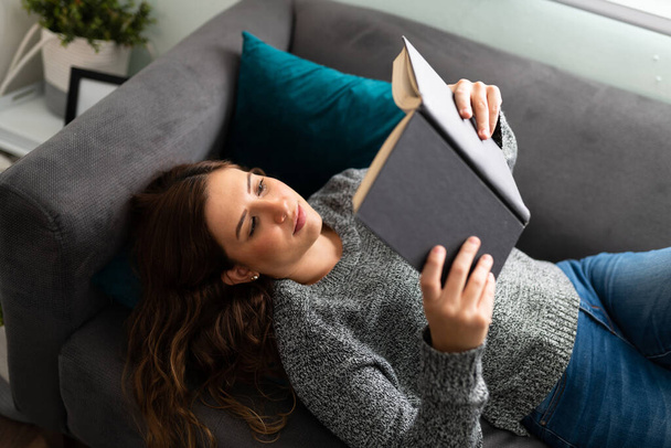 Atraktivní žena po dvacítce spatřena shora, ležící na pohovce v obývacím pokoji a čtoucí knihu - Fotografie, Obrázek