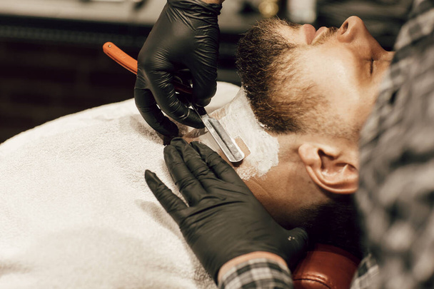 Barber shaving a bearded man in a barber shop. Close up of a hairdresser's hands in black gloves shaving a client's beard. - Фото, зображення