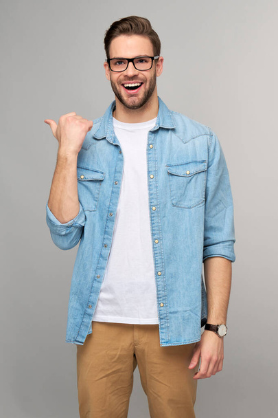 Šťastný mladý pohledný muž v džínách košile ukazuje pryč stojící proti šedému pozadí - Fotografie, Obrázek