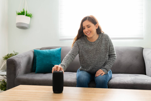 Lächelnde Frau in ihren Zwanzigern schaltet ihren neuen smarten Lautsprecher in ihrem Wohnzimmer ein  - Foto, Bild