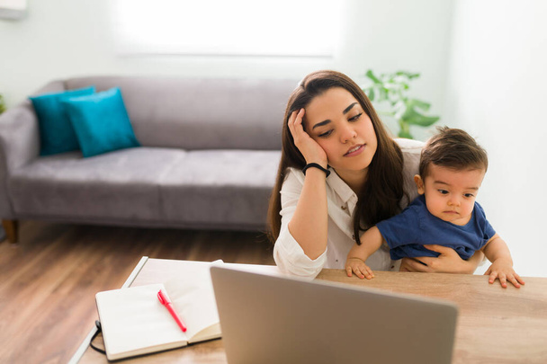 Evde dizüstü bilgisayarla uğraşırken bebekle masada oturan kadın yorgun görünüyor. - Fotoğraf, Görsel
