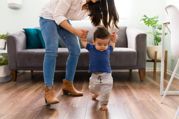 Adorable baby boy taking his first steps on wooden floor with help of his mother at home - Zdjęcie, obraz