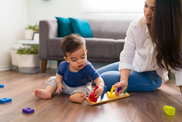 Adorable baby boy sitting on floor playing with educational toys, arranging and sorting colors and shapes while her mother helping him - Фото, изображение