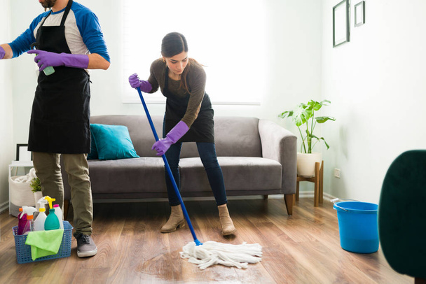 Przystojna kobieta myje podłogę, podczas gdy jej chłopak odkurza i czyści meble w salonie - Zdjęcie, obraz