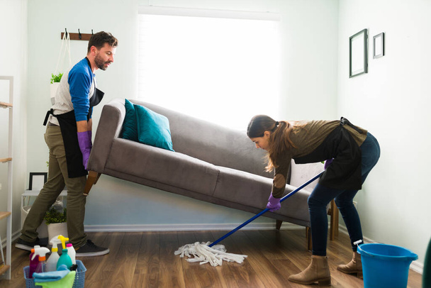 Mann hebt die Couch im Wohnzimmer und seine Frau wischt unter ihr, während sie gemeinsam die Wohnung putzt - Foto, Bild