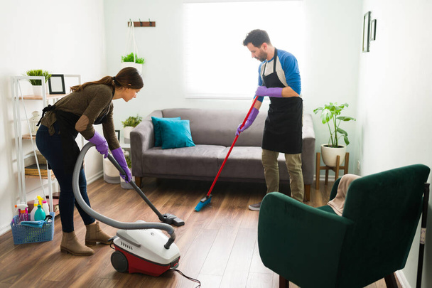 Vollständige Ansicht eines Paares mit einem Besen und einem Staubsauger, um den Wohnzimmerboden zu Hause zu reinigen - Foto, Bild