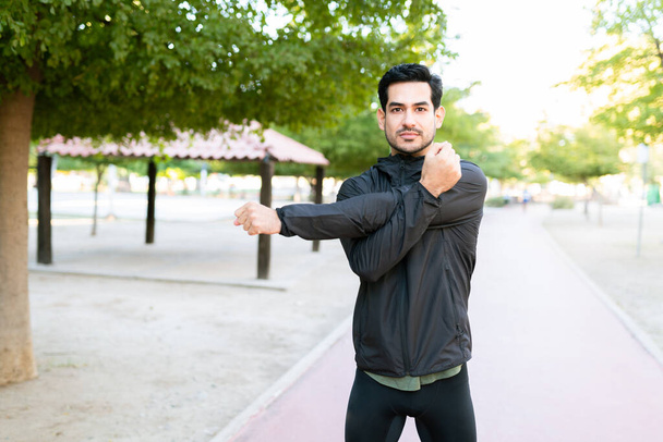 Uomo attraente sulla ventina in abbigliamento sportivo che allunga i muscoli del braccio prima del suo allenamento cardio ed esercizi nel parco  - Foto, immagini
