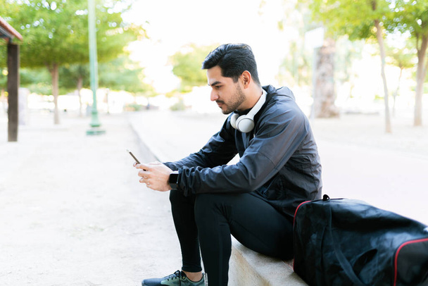 A latin férfi sms-ezik a mobilján a parkban végzett gyakorlata után. Egy férfi sportruházatban pihen a fülhallgatójával és egy sporttáskával.  - Fotó, kép