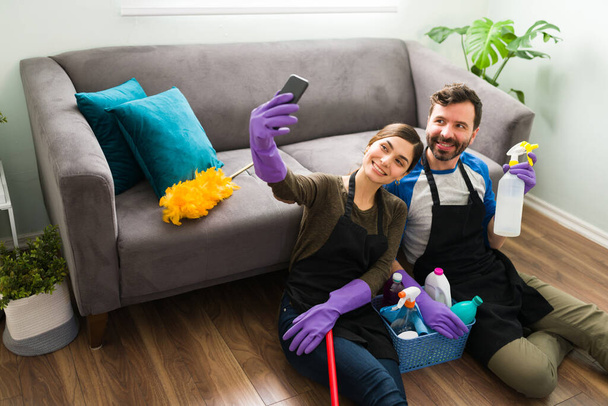 Pareja hispana guapa usando guantes y un delantal tomando una selfie mientras limpia y hace trabajo en casa - Foto, Imagen