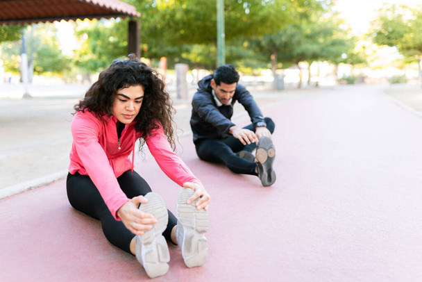 Atlétikai pár kinyújtóztatja a lábát a futópályán, mielőtt futni menne. Latin pár csinál egy ülő előre kanyar - Fotó, kép