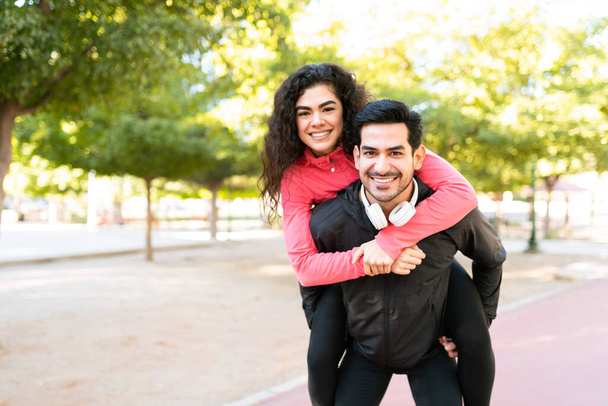 Portré egy fiatal spanyol párról, akik sportos ruhákat viselnek a parkban. Vonzó latin férfi, aki a csinos barátnőjét cipeli a hátán.  - Fotó, kép