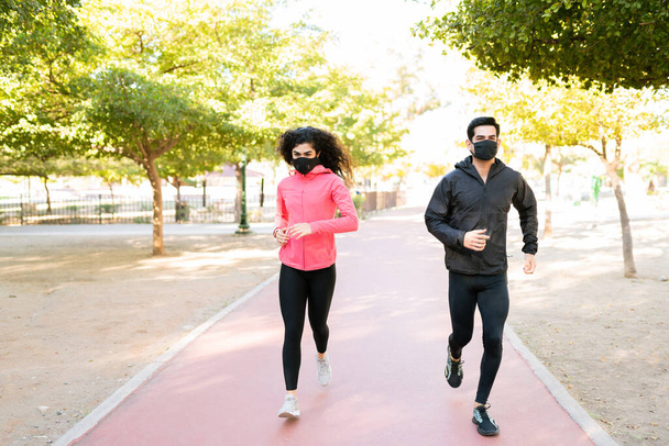 Hombre joven y mujer joven con un estilo de vida saludable corriendo al aire libre durante la pandemia del coronavirus. Ajuste pareja con máscaras faciales y trotar en el parque - Foto, Imagen