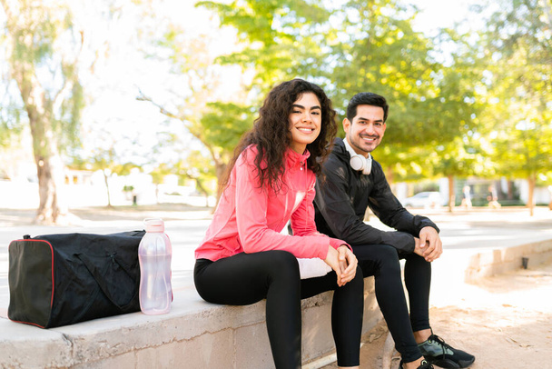 Çekici genç bir kadın ve elinde su şişesi ve spor çantasıyla gülümseyen yakışıklı bir adam. Spor kıyafetleri giyen ve antrenmandan sonra parkta dinlenen bir çift.  - Fotoğraf, Görsel