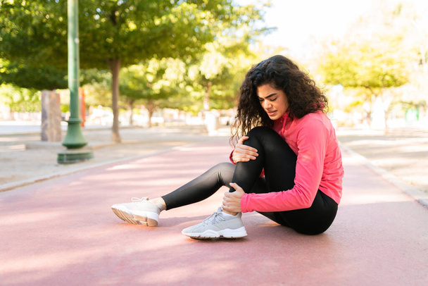 Ισπανίδα νεαρή γυναίκα στα αθλητικά υποφέρει από τραυματισμό στο πόδι στην πίστα του πάρκου. - Φωτογραφία, εικόνα