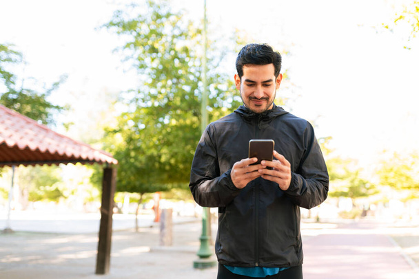 Latino komea mies hänen 20s yllään urheilullinen vaatteita tekstailee hänen älypuhelin puistossa - Valokuva, kuva
