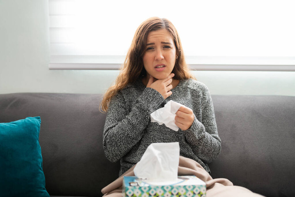 Young woman suffering from throat pain and a cough. Portrait of sick woman sitting on the sofa with a box of tissues - Photo, Image