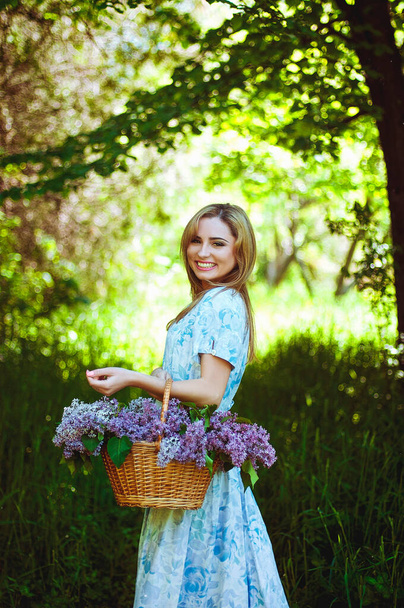 Portrait of young woman in blue dress in spring garden with a basket full of lilacs. Spring background. Woman's Day. Greeting card.  - Zdjęcie, obraz