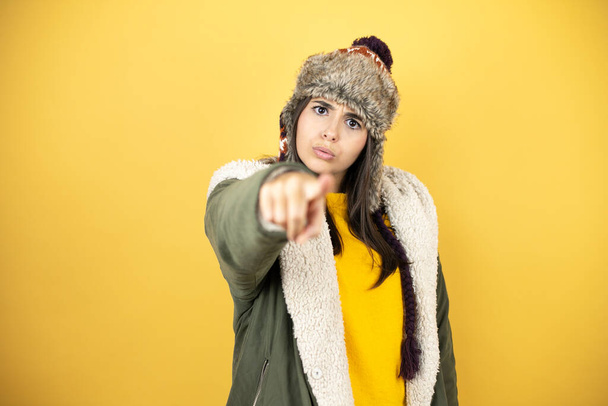 Sarı arkaplan üzerine yeşil bir palto ve şapka takan genç ve güzel bir kadın ön tarafı parmağıyla işaret ediyor. - Fotoğraf, Görsel