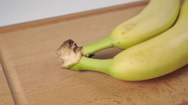 Banany zbliżają się do dwóch nieobranych egzotycznych owoców z zieloną łodygą na drewnianej desce do krojenia - Materiał filmowy, wideo