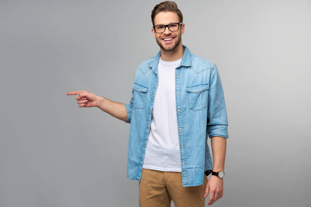 Happy young handsome man in jeans shirt pointing away standing against grey background - Foto, Bild