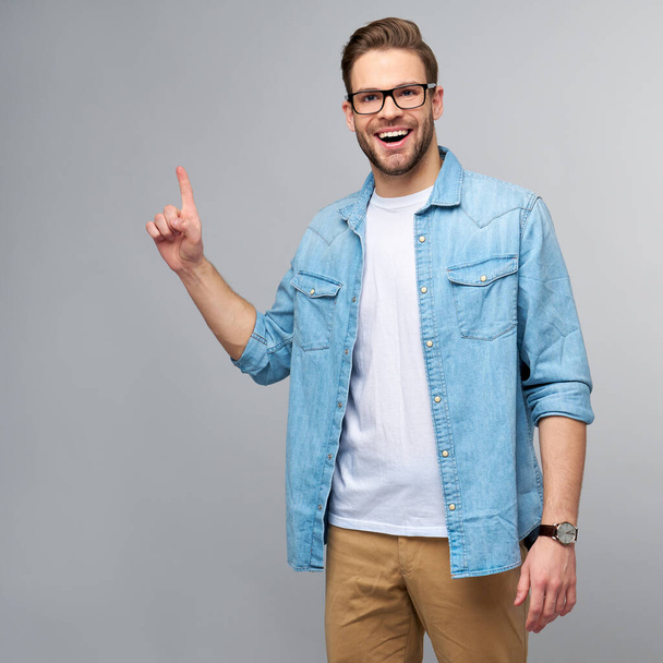 Happy young handsome man in jeans shirt pointing away standing against grey background - Fotografie, Obrázek