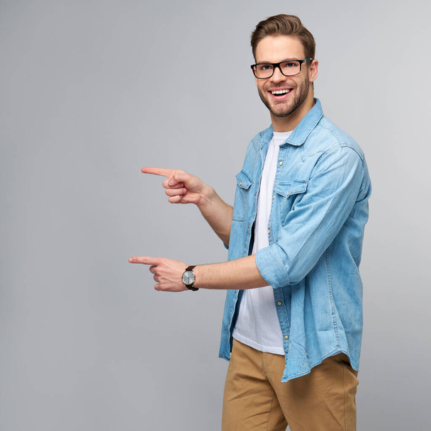 Happy young handsome man in jeans shirt pointing away standing against grey background - Foto, immagini