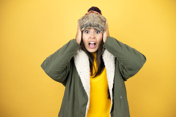 Jonge mooie vrouw met een hoed en een groene winterjas over gele achtergrond gek en bang met handen op het hoofd, bang en verrast van shock - Foto, afbeelding