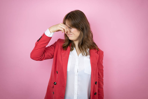 Young business woman over isolated pink background smelling something stinky and disgusting, intolerable smell, holding breath with fingers on nose - Photo, Image