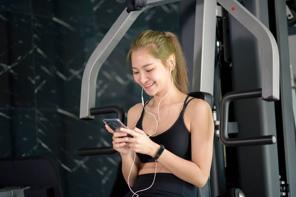 A beautiful smiling woman in earphones using a social network and listening to music at gym, Fitness and technology concept - Фото, изображение