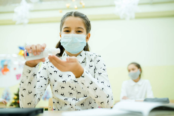 Ein Mädchen sitzt in einer Klasse bei einer Unterrichtsstunde in einer medizinischen Maske und benutzt ein Antispetikum für die Hände - Foto, Bild