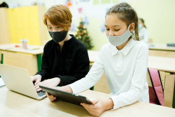 Students in medical masks sit at the table and take part in an online lesson using gadgets - Photo, Image