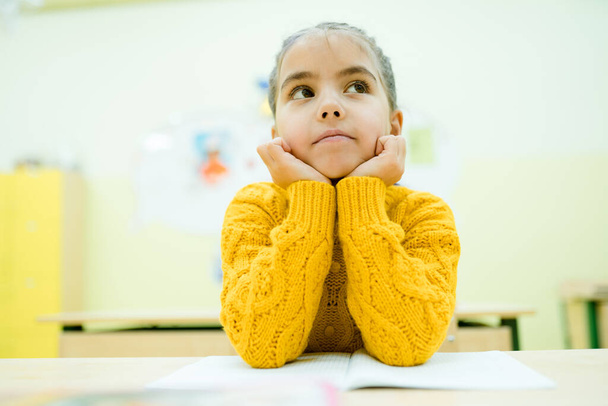 Sarı örgü süveterli küçük bir çocuk, dirsekleri elinde okul masasında oturuyor. - Fotoğraf, Görsel