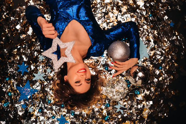 a woman in a blue sequined dress smiles and lies on the floor under a falling multicolored confetti - Photo, Image
