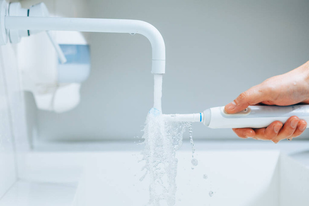 рука держит зубную щетку под струей воды из крана в ванной комнате - Фото, изображение