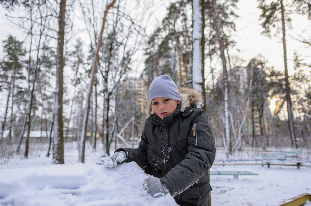 leuke witte jongen in een warme jas en hoed speelt met sneeuw op de speelplaats in het park in de winter - Foto, afbeelding