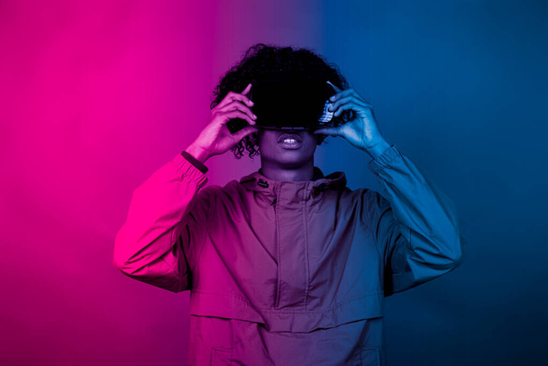 VRメガネをかけたアフロボーイ。仮想現実に没頭する若いアフロが、手で動き出す。青と赤の光が少年に降りかかる. - 写真・画像