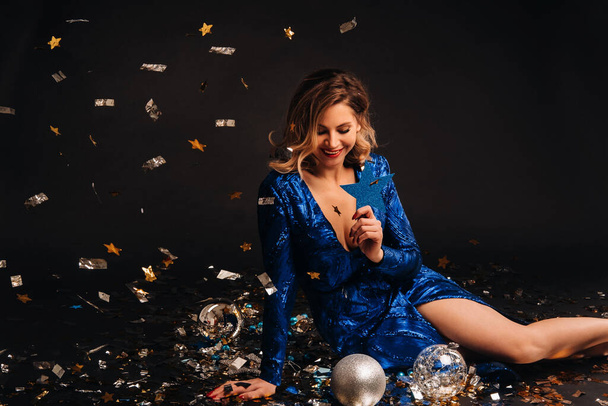 a woman in a blue sequined dress is smiling sitting on the floor with confetti on a black background. - Photo, Image