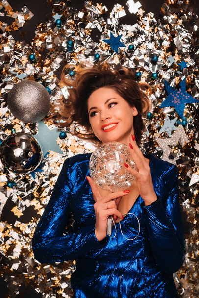 a woman in a blue sequined dress smiles and lies on the floor under a falling multicolored confetti - Foto, Imagen