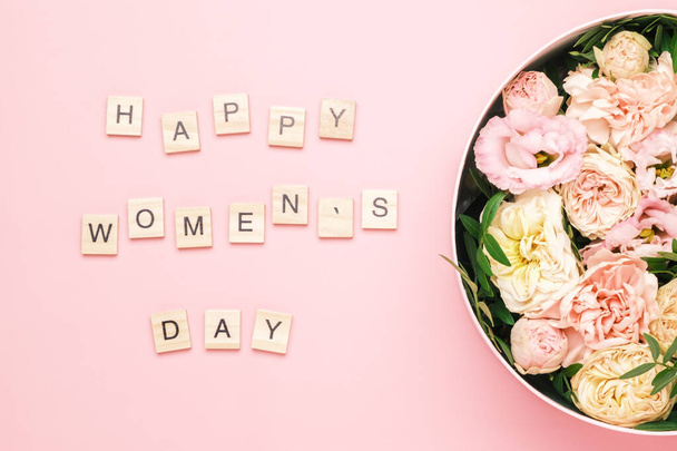 Happy womens day lettering on the wooden squares with letters on the pink background and big round box with red flowers and roses. - Photo, Image