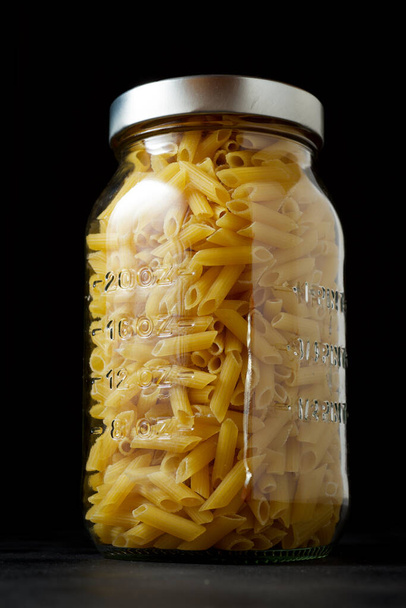Glass jar full of macaroni, on a black table. - Фото, зображення