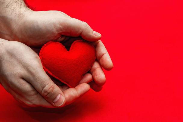 Η ιδέα του Αγίου Βαλεντίνου. Τα αρσενικά χέρια κρατούν μια σπιτική καρδιά από τσόχα. Φωτεινό κόκκινο ανάγλυφο φόντο - Φωτογραφία, εικόνα