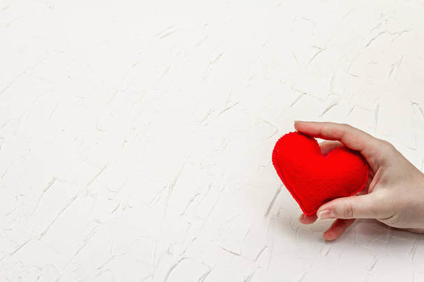 Η ιδέα του Αγίου Βαλεντίνου. Το γυναικείο χέρι κρατάει μια σπιτική κόκκινη καρδιά από τσόχα. Άσπρο στόκο ανάγλυφο φόντο - Φωτογραφία, εικόνα