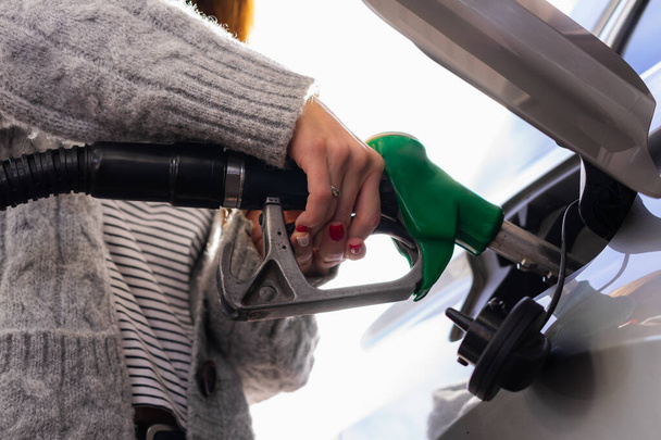Nuori nainen, jolla on maalatut kynnet ja mukava pusero, täyttää autonsa bensiinillä auttamalla itseään kahdella kädellä. Matalasta kulmasta otettu kuva. - Valokuva, kuva