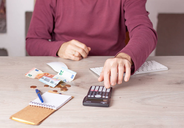 Las manos de un hombre en una mesa de oficina haciendo cálculos económicos con una calculadora más dinero un bloc de notas. - Foto, Imagen